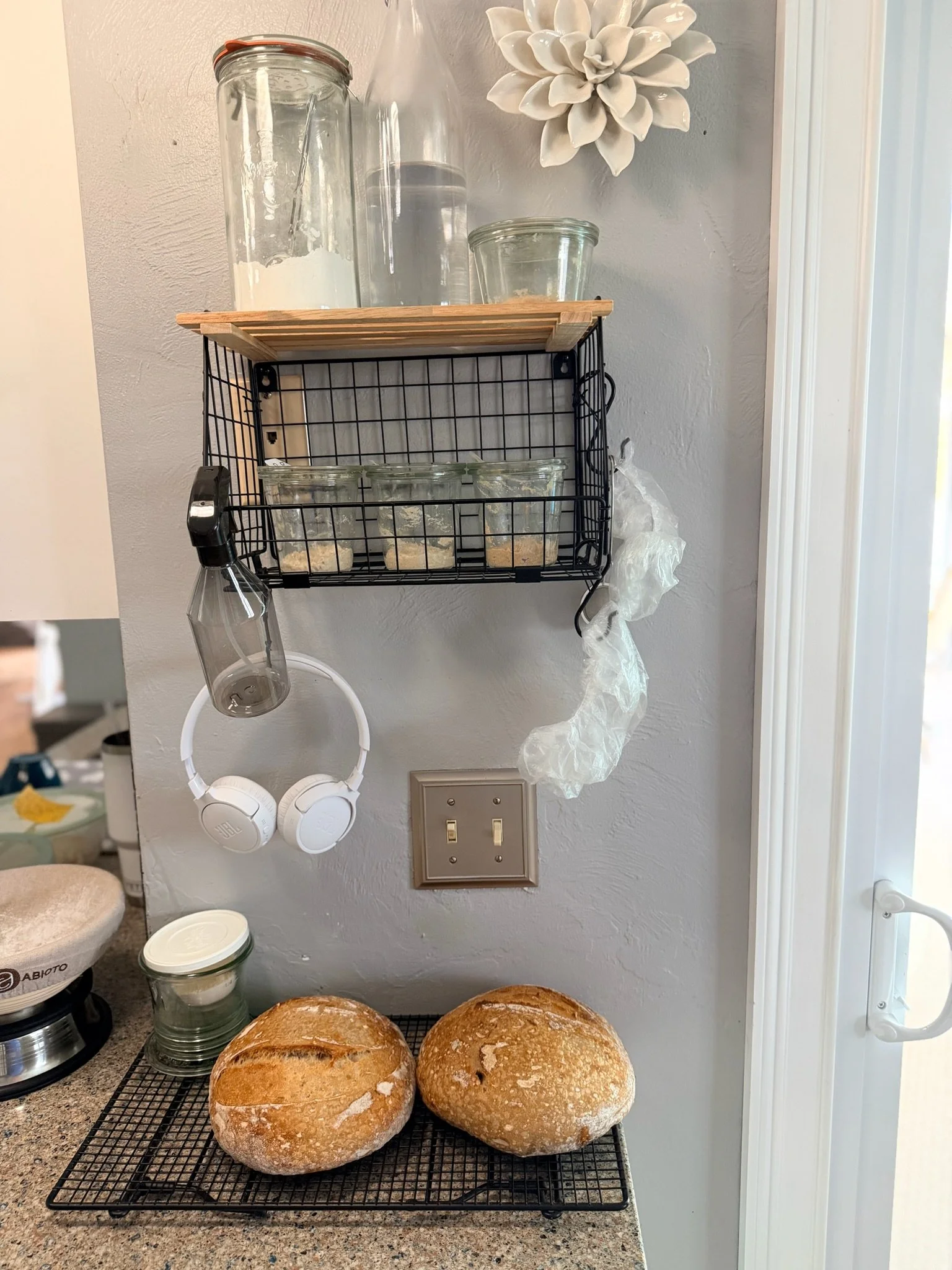 Three loaves of freshly baked bread on a cooling rack in a kitchen. Above, a wall-mounted black wire shelf holds glass jars and containers, with a decorative white flower on the wall. A pair of white headphones hang below the shelf, and a coffee make