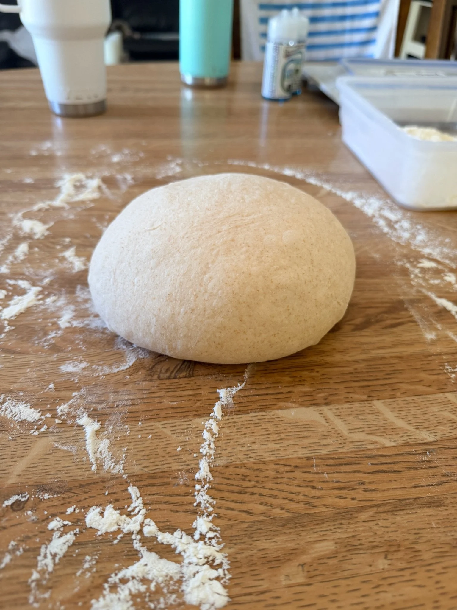 A ball of pizza dough on a wooden table, with scattered flour around it and kitchen items in the background.