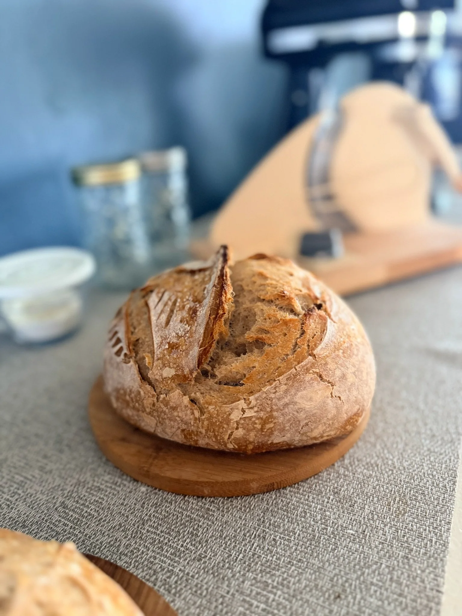 A rustic baguette bread on a wooden cutting board placed on a gray fabric surface with a kitchen appliance in the background.