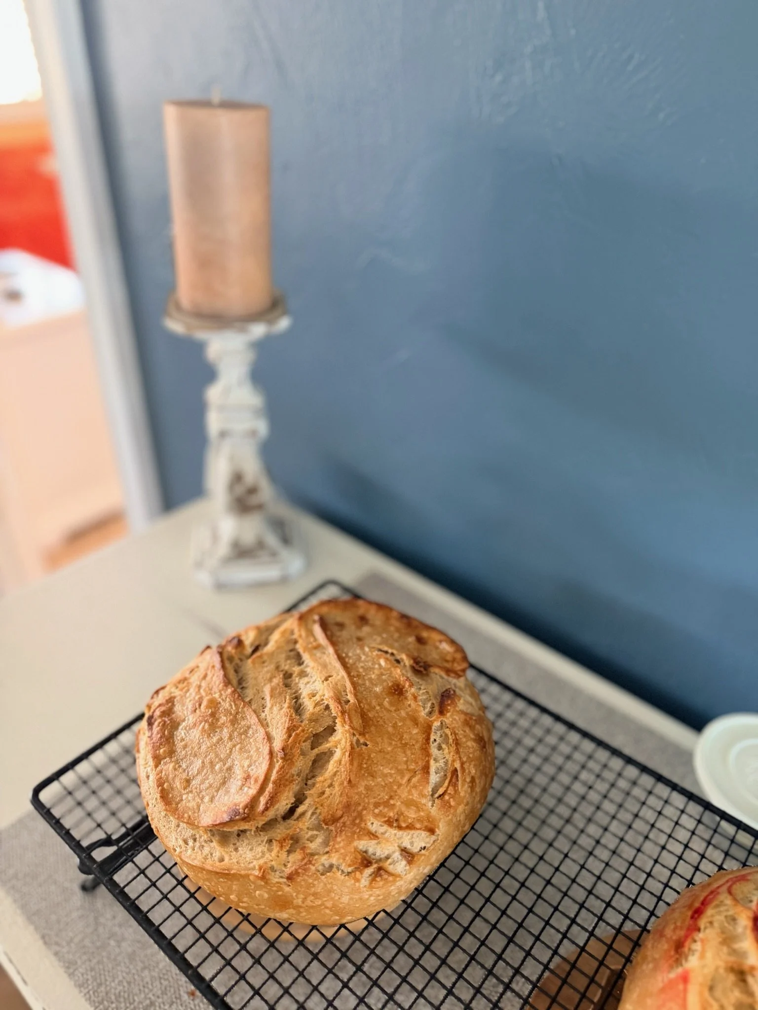 A loaf of freshly baked bread with a golden crust, cooling on a black wire rack, against a blue wall.