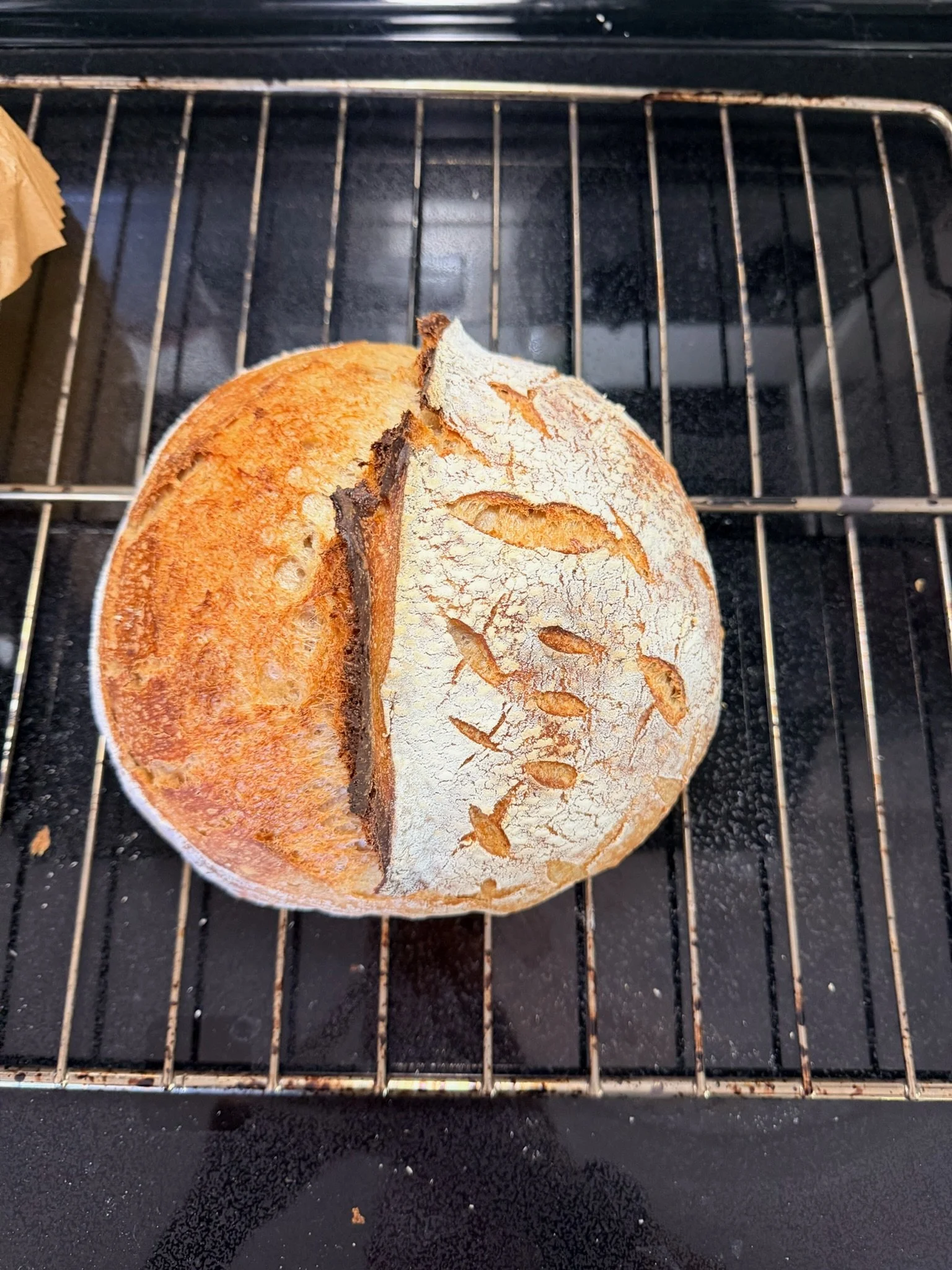 A loaf of bread with a crack on top is cooling on a wire rack, placed on a black countertop.