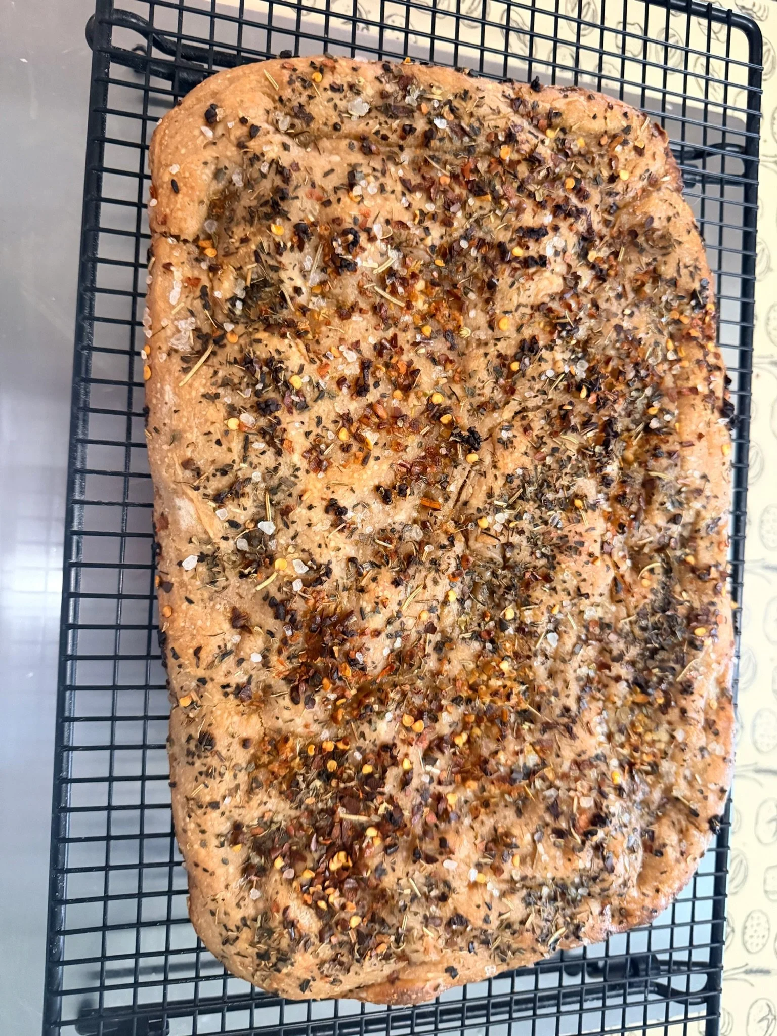 Three loaves of bread topped with herbs and spices, cooling on a black wire rack.