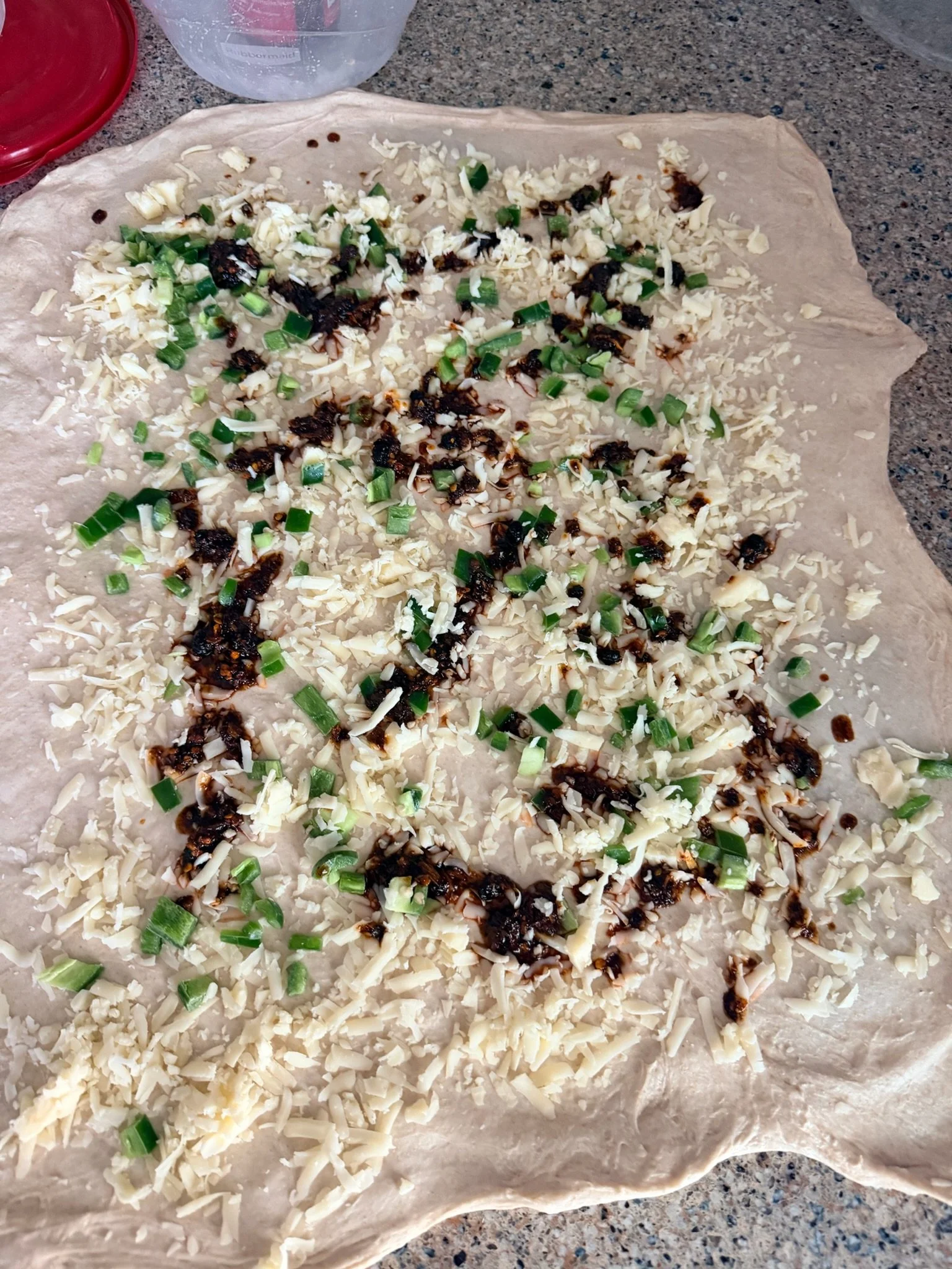 Unrolled dough topped with shredded cheese, chopped green onions, and a dark sauce on a parchment paper on a countertop.