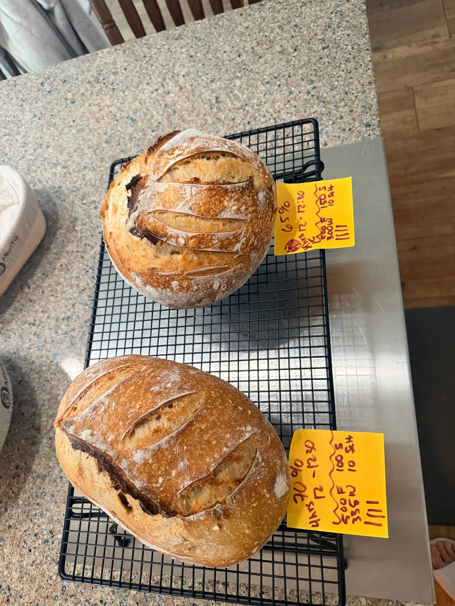 A round loaf of bread with a crusty, golden-brown exterior and decorative shallow slashes on top, placed on a black wire cooling rack. Two yellow handwritten price tags are positioned beside it on a speckled granite countertop.