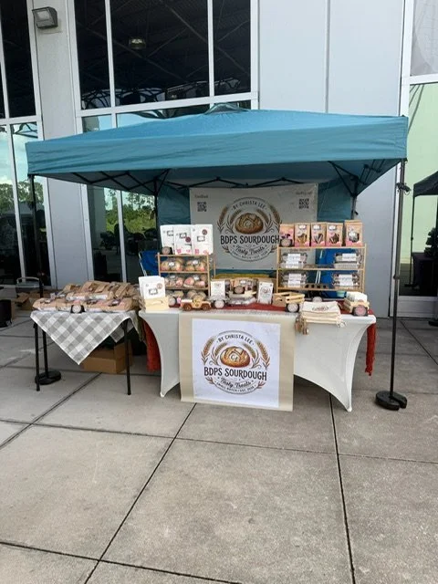 Market stall selling sourdough bread and baked goods, set up under a blue canopy outside a building.