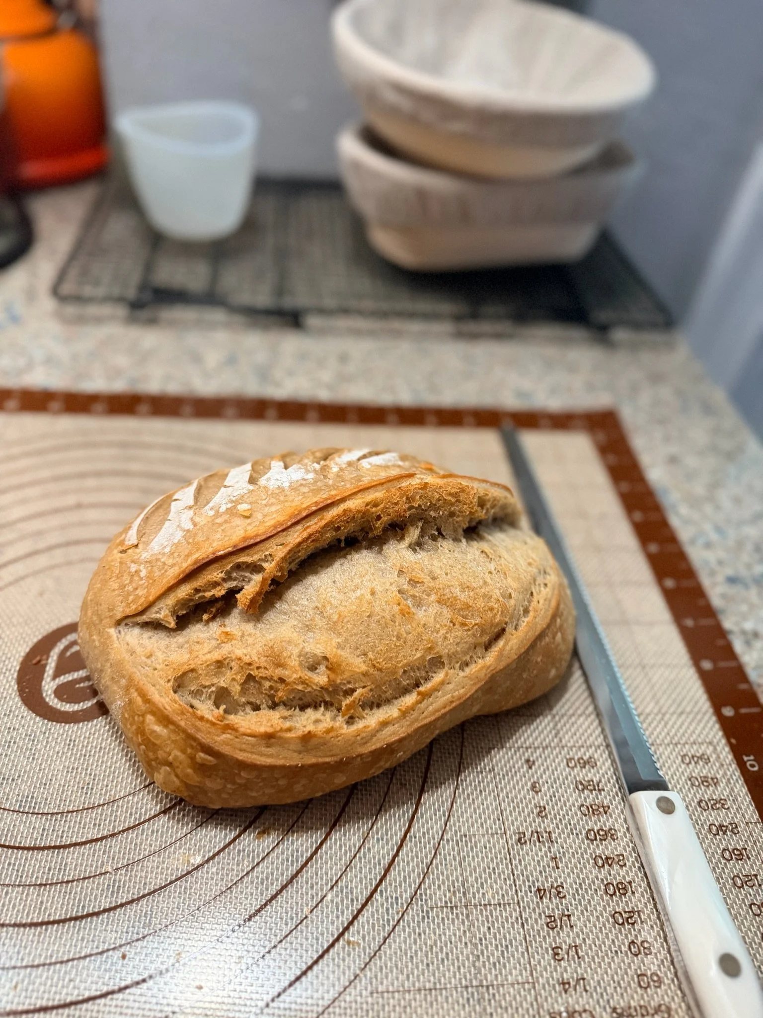 A partially sliced loaf of bread on a silicone baking mat with a measuring kit and a thermometer nearby, in a kitchen setting.