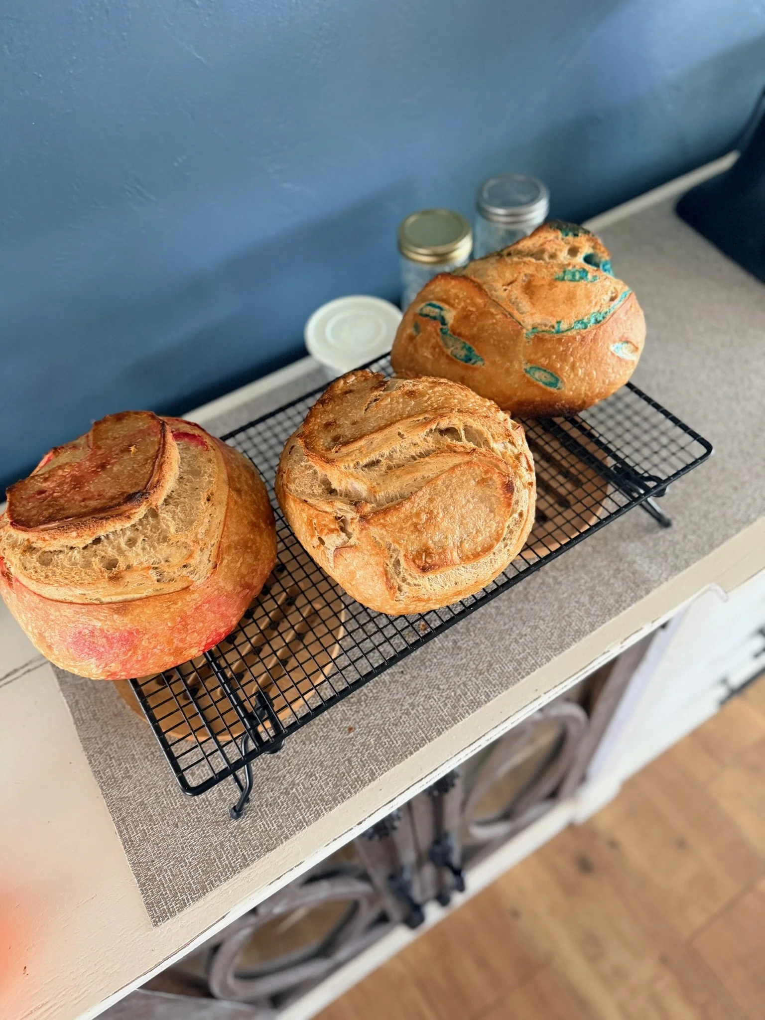 Three freshly baked bread loaves with golden crusts on a black cooling rack, placed on a beige cloth on a white table.