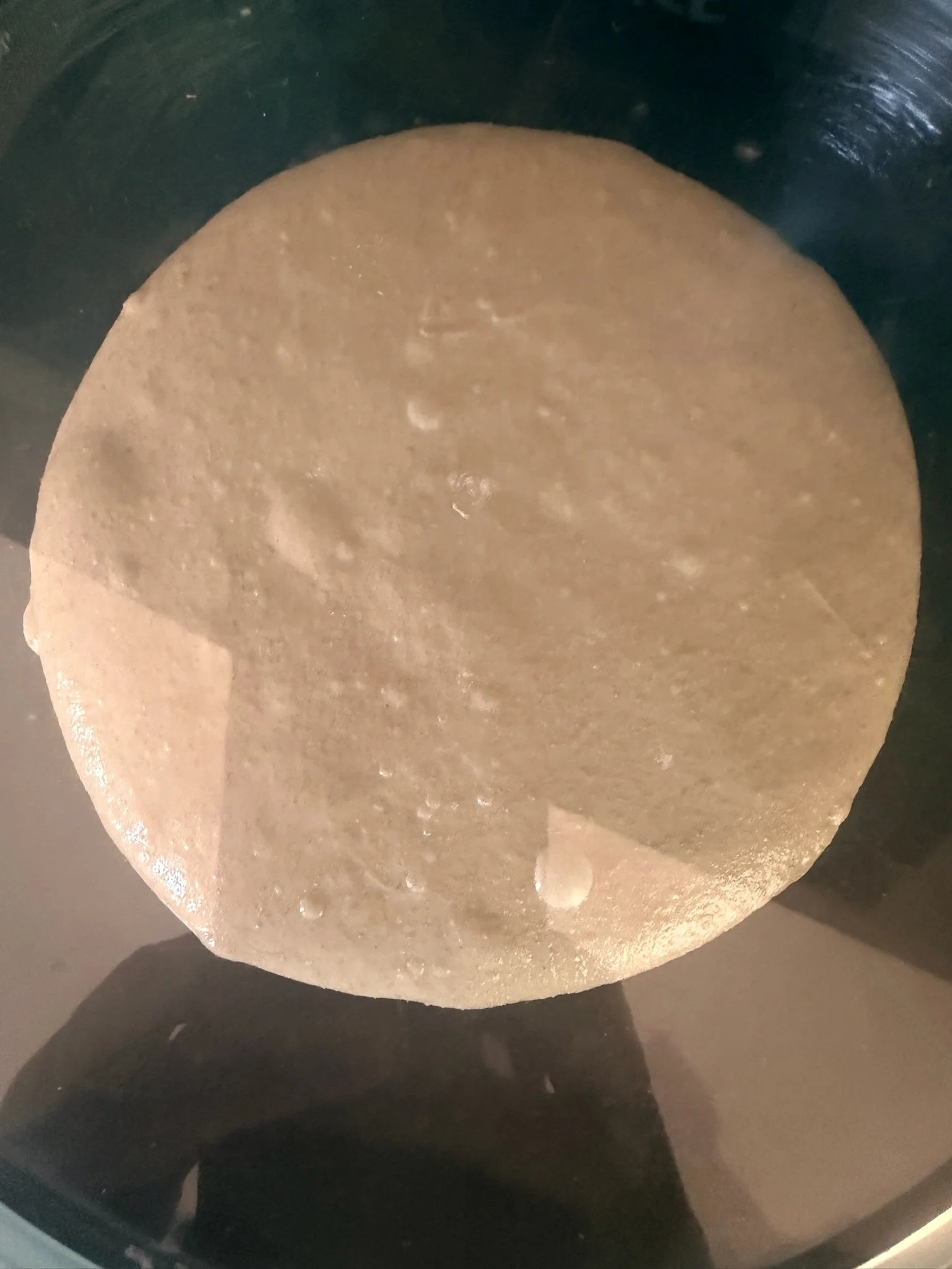 A round, flat piece of dough, possibly for making bread or pizza, resting on a black surface, likely a baking stone or pan.
