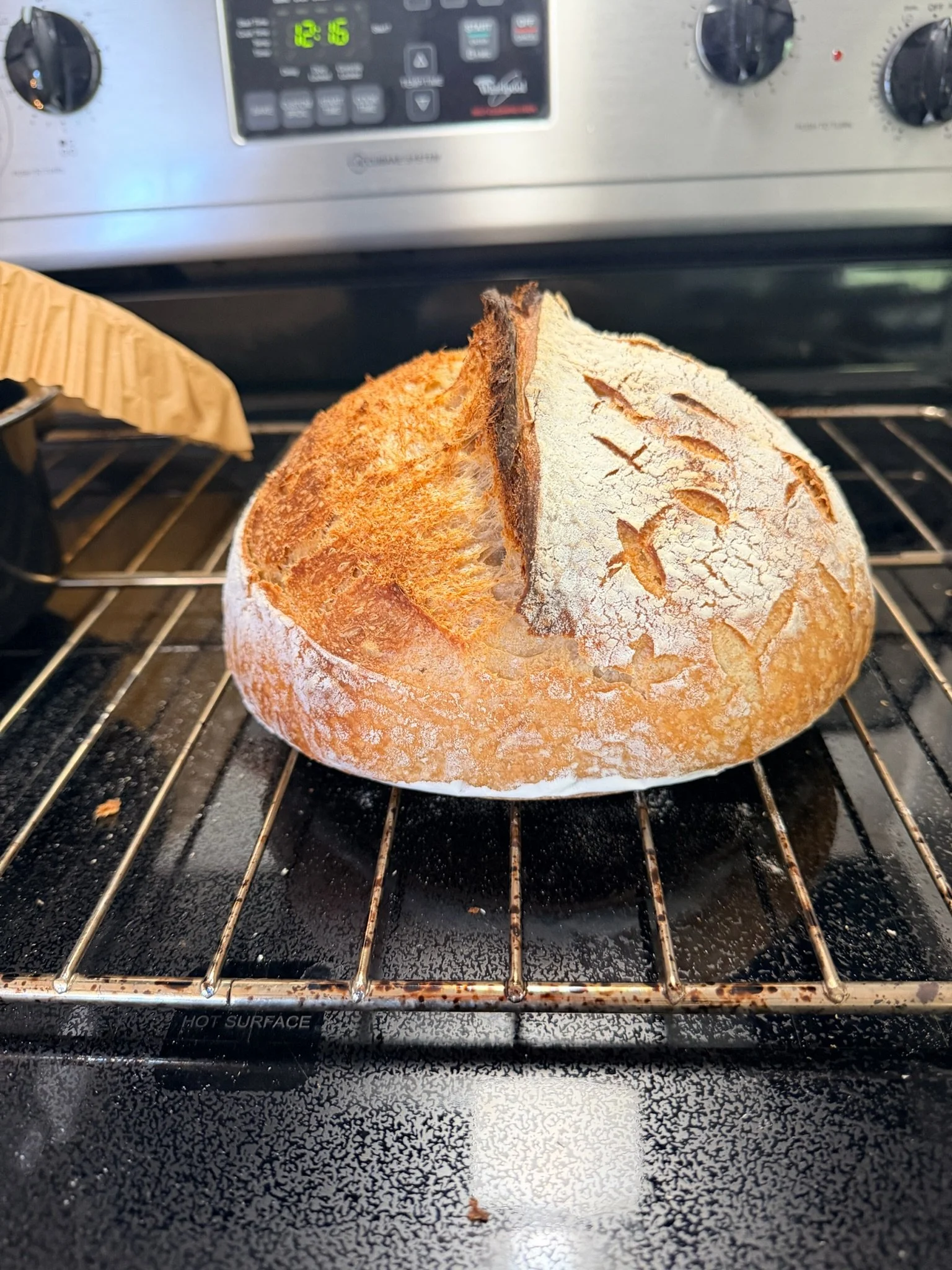 A round loaf of bread with a crusty golden exterior and decorative scoring on top, resting on an oven rack inside a kitchen oven.