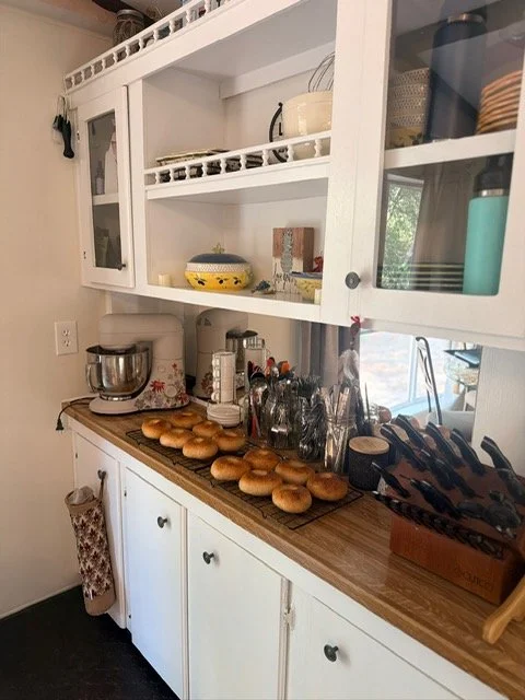 Kitchen counter with a tray of bagels, coffee making equipment, and utensils, with white cabinets and a window in the background.