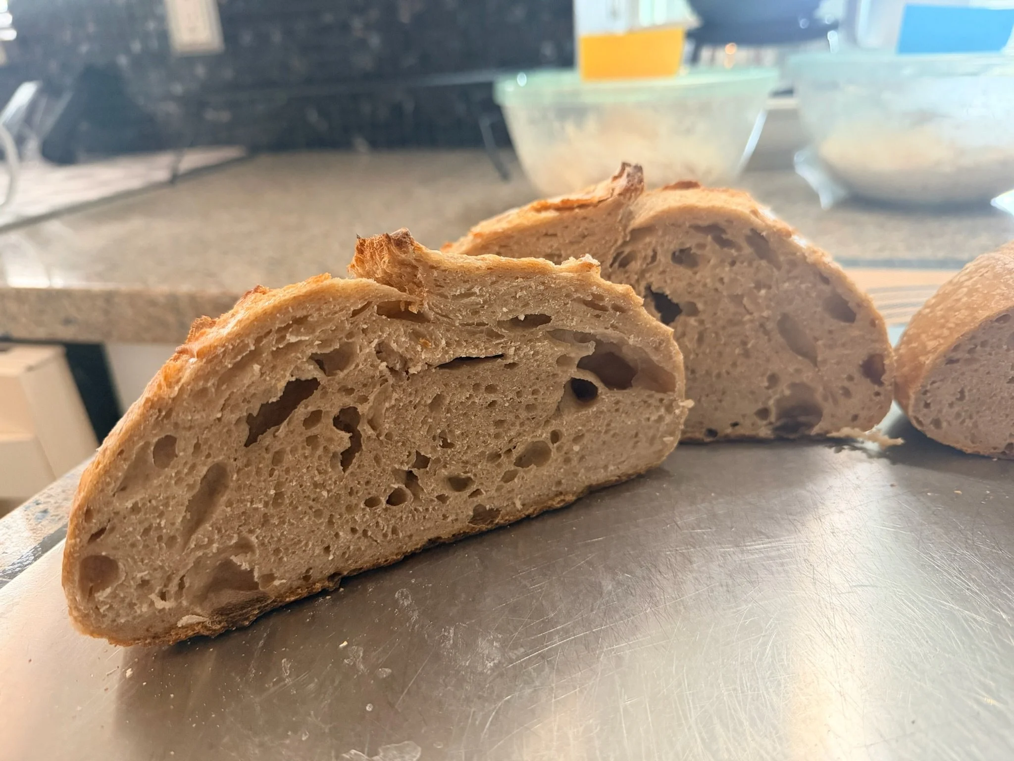A loaf of bread cut into two pieces, showing a light brown crust and a soft interior with irregular holes, resting on a metal surface in a kitchen.