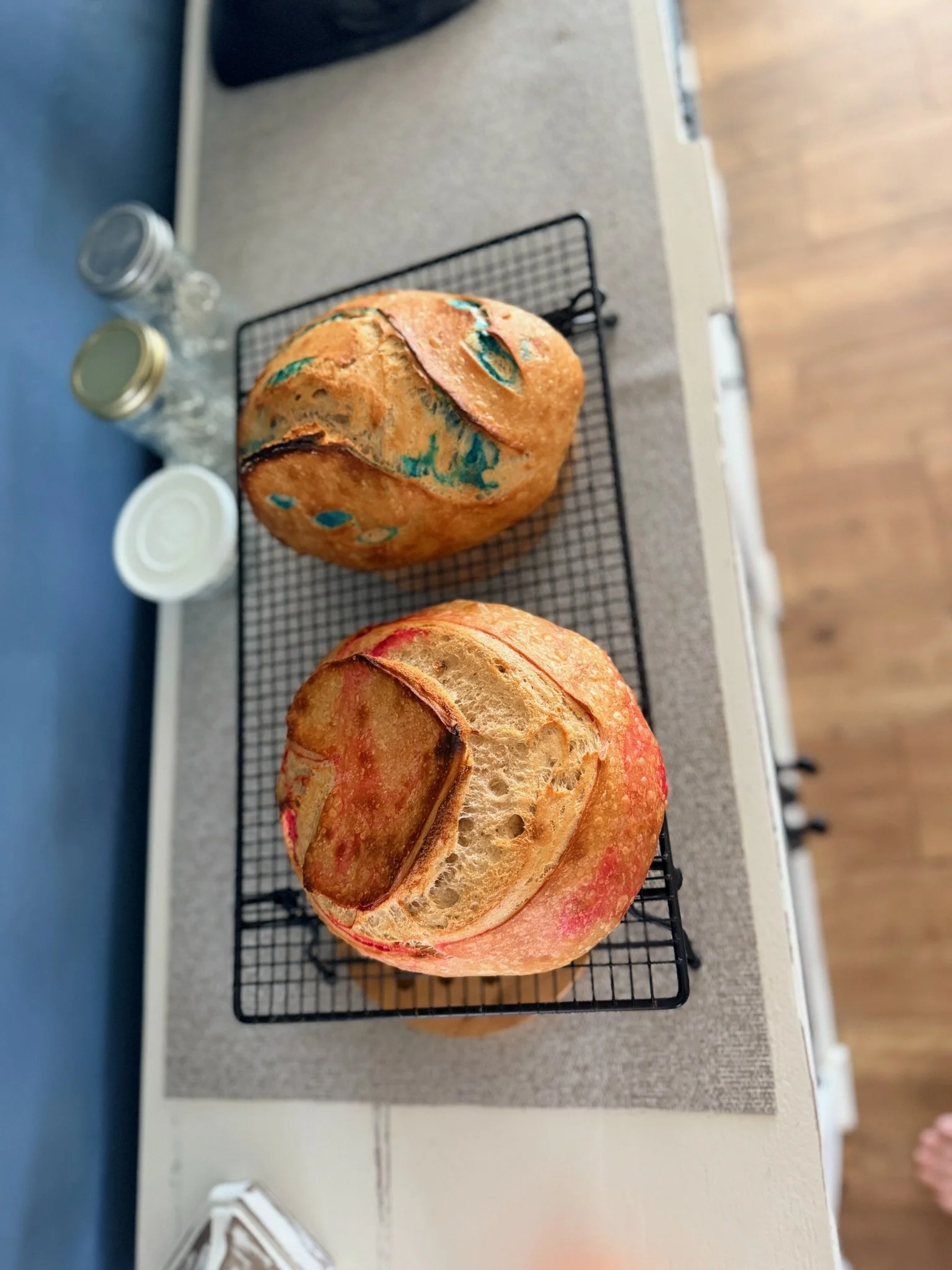 Two freshly baked loaves of bread, one with a reddish crust and the other with a golden crust, resting on a wire cooling rack on a kitchen countertop.