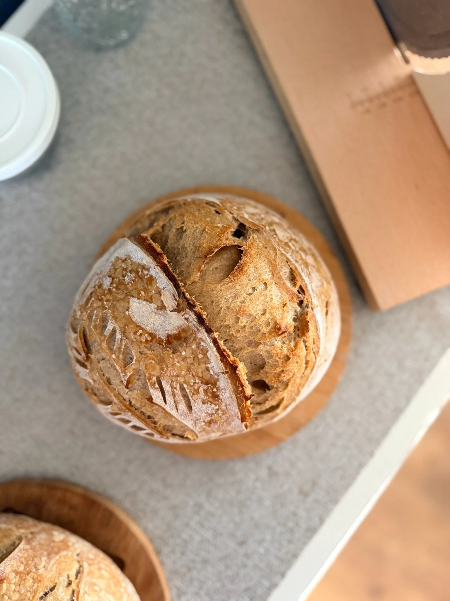 Close-up of a rustic loaf of bread on a wooden cutting board with or without a knife, beside a terracotta bowl and a white container, on a gray countertop.