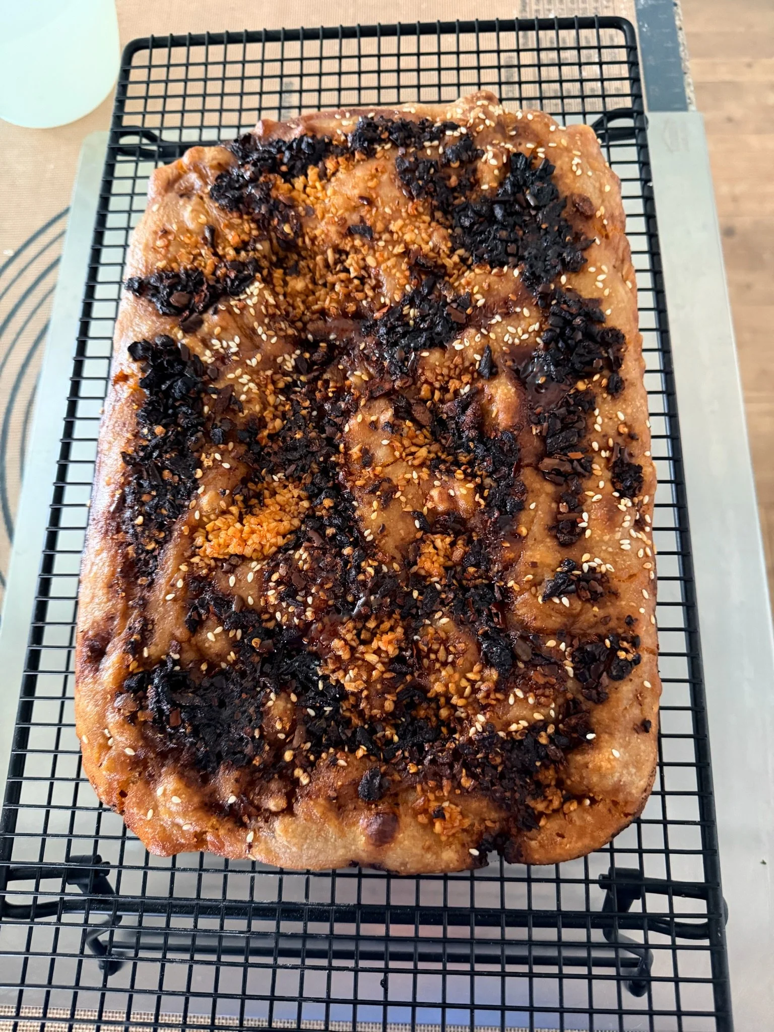 A rectangular loaf of bread topped with various crushed nuts and seeds, some of which are dark and some light, on a cooling rack.