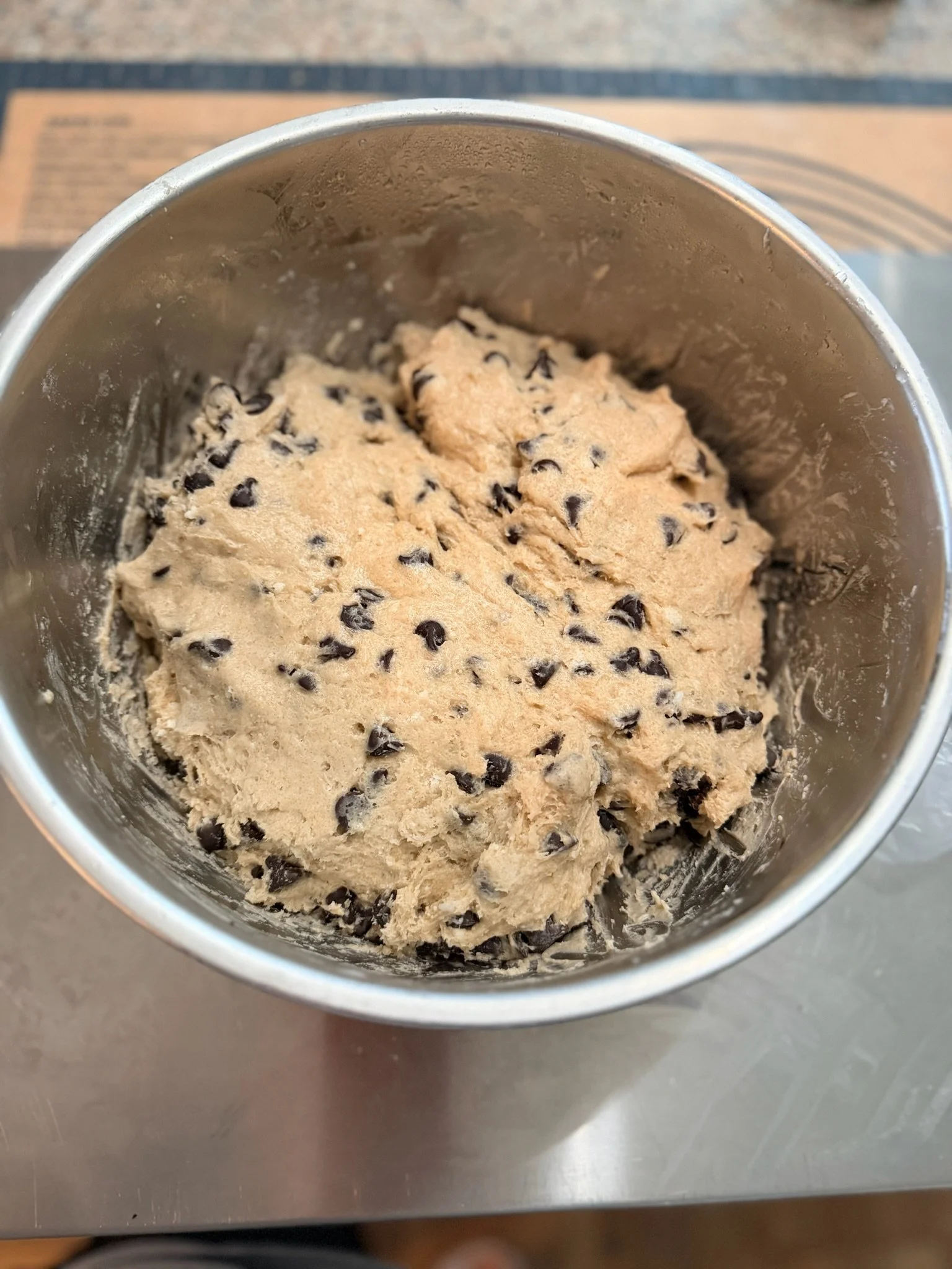 Close-up of cookie dough with chocolate chips in a metal mixing bowl on a stainless steel countertop.