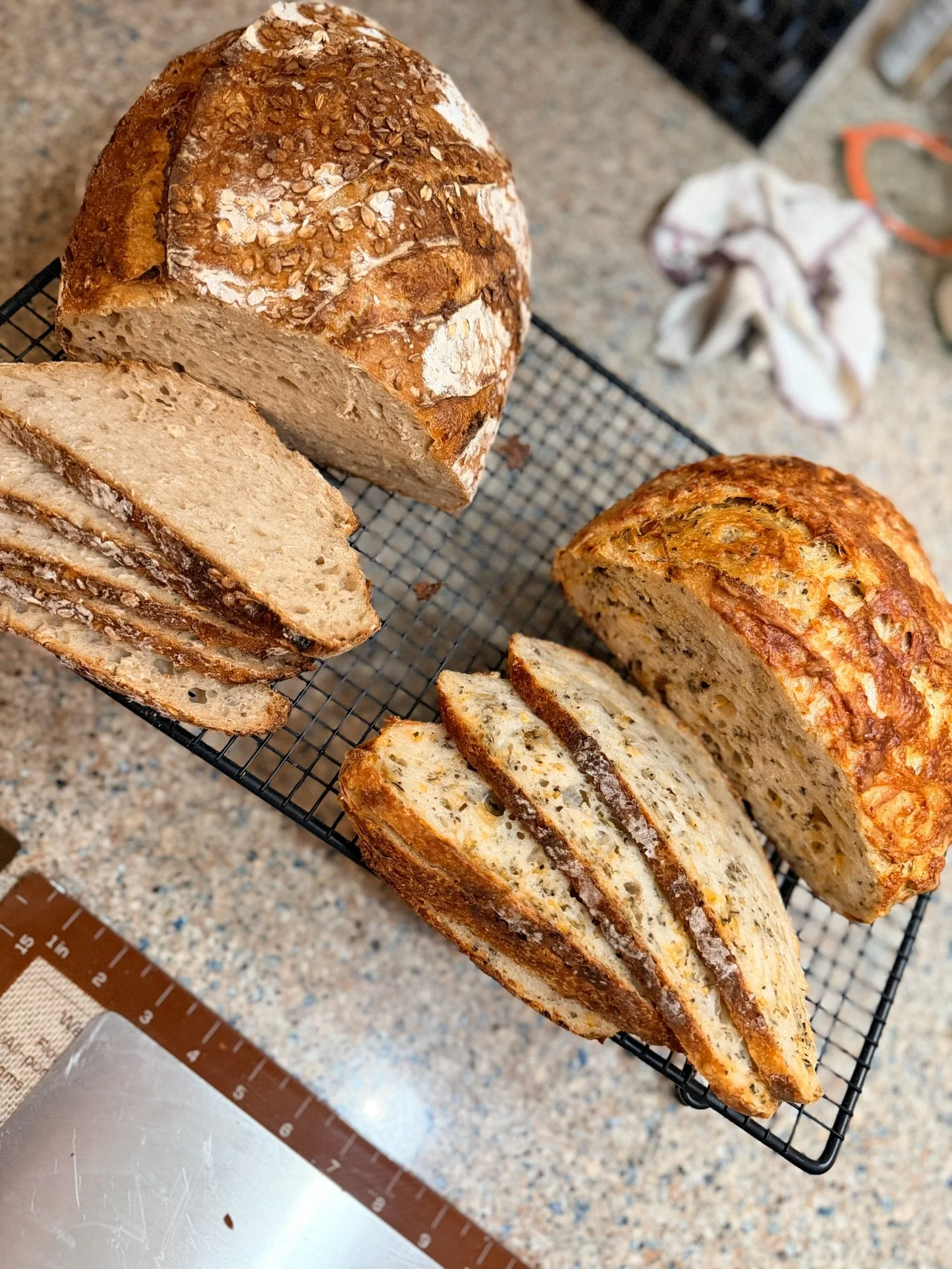 A loaf of bread on a cooling rack, sliced to show the interior, with a golden crust topped with oats. The background includes a speckled countertop and a cloth.