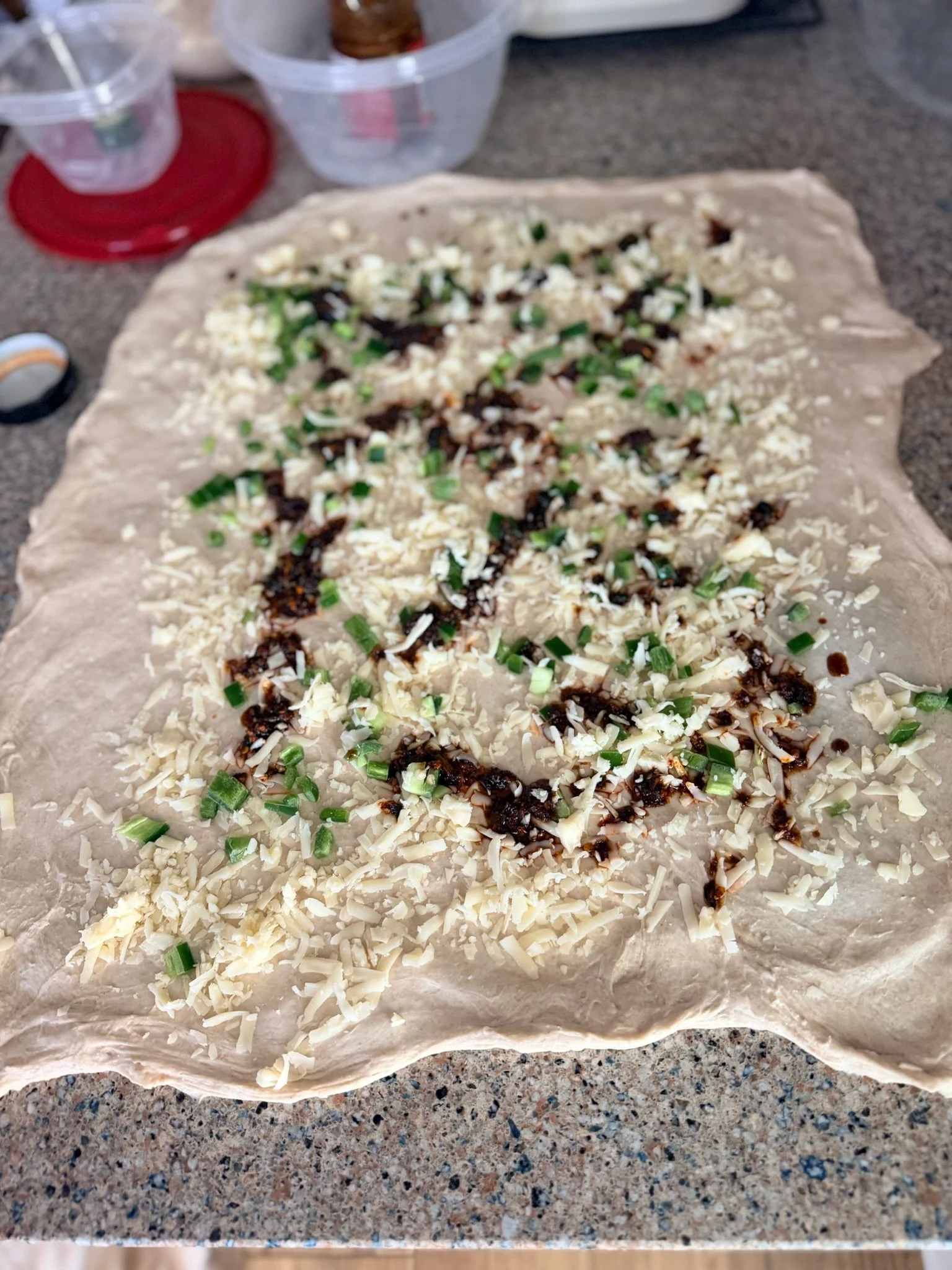 Unrolled pastry dough topped with shredded cheese, chopped green onions, and drizzled with a dark sauce, on a kitchen countertop.