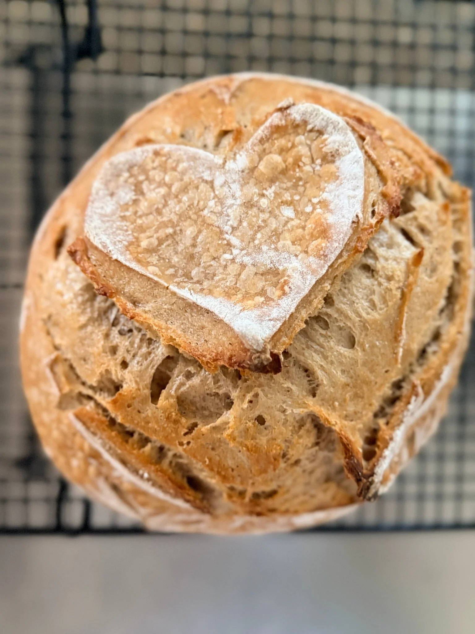 A close-up of bread with a coarse texture, featuring a white heart-shaped pattern dusted with flour on top.