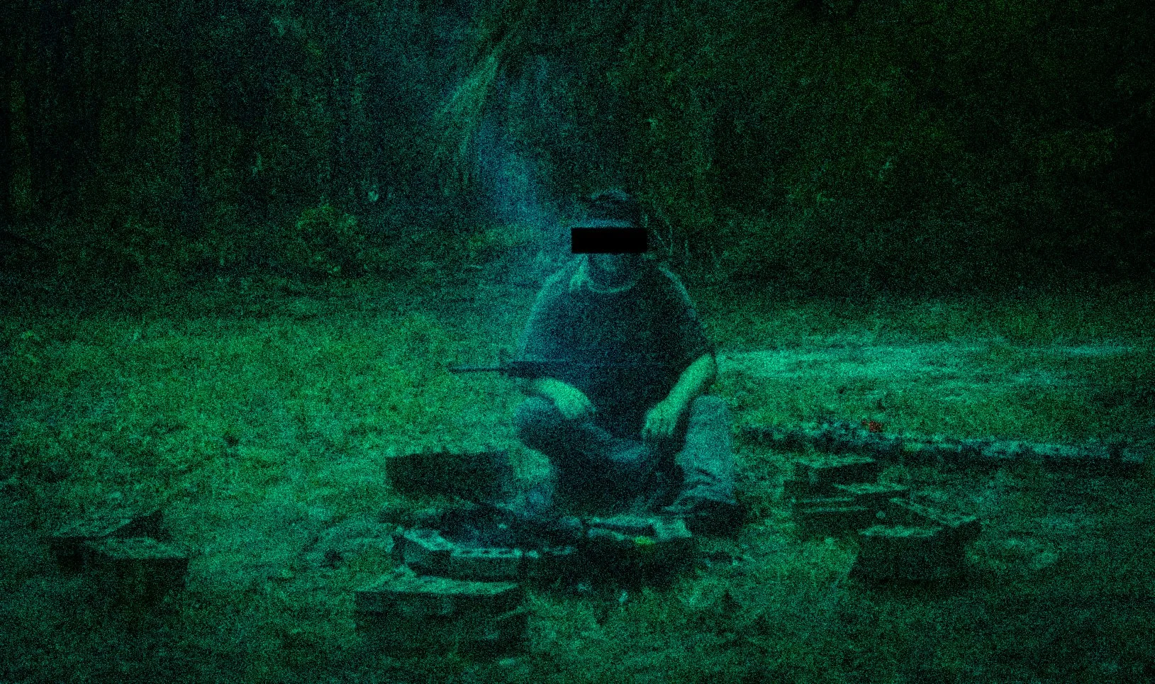 A person, sitting cross-legged outdoors on a blanket, surrounded by scattered objects, under a tree during nighttime.