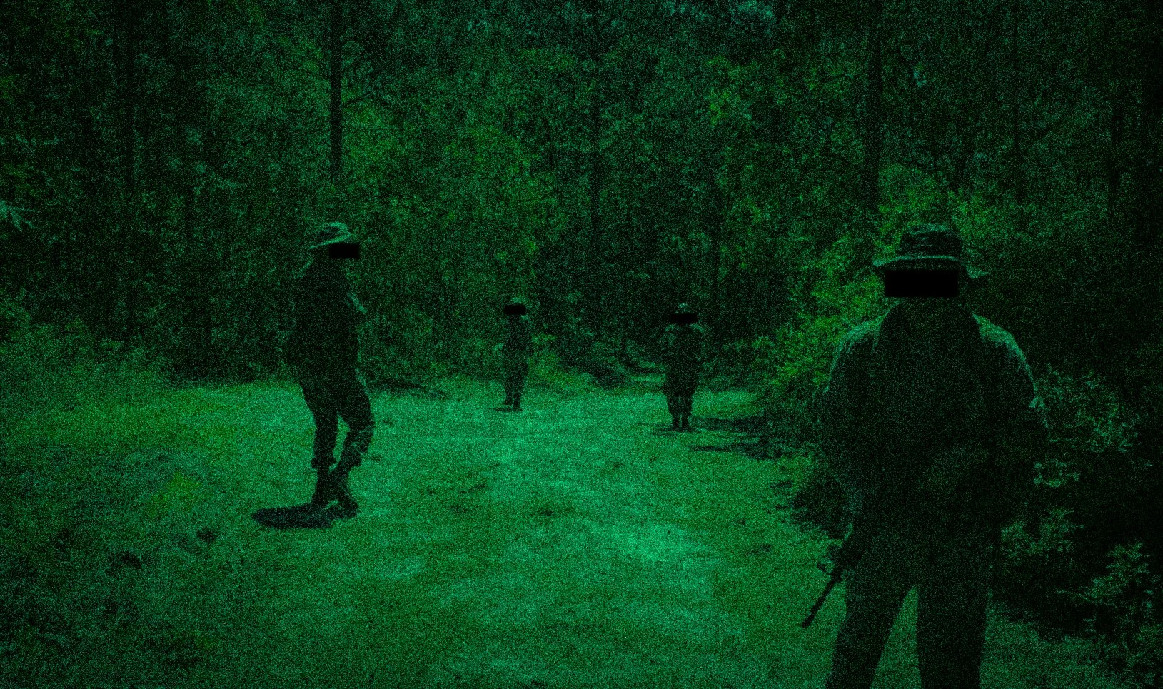 Four airsoft operators walking along a wooded trail at night, illuminated by green night vision.