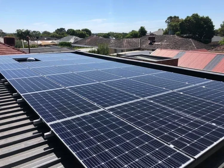 Solar panels installed on a rooftop, with a neighborhood and trees in the background under a clear blue sky.