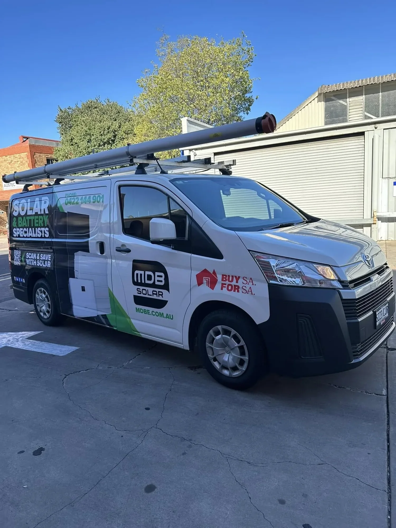 A white company van with solar and battery service advertisements parked on cracked pavement in front of a warehouse building. The van has a long aerial solar panel on top and a prominent logo on the side.