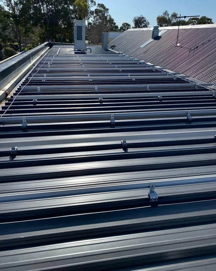 Metal roof with solar panel mounting rails and a solar panel installed at the top, with trees and part of a neighboring roof visible in the background.