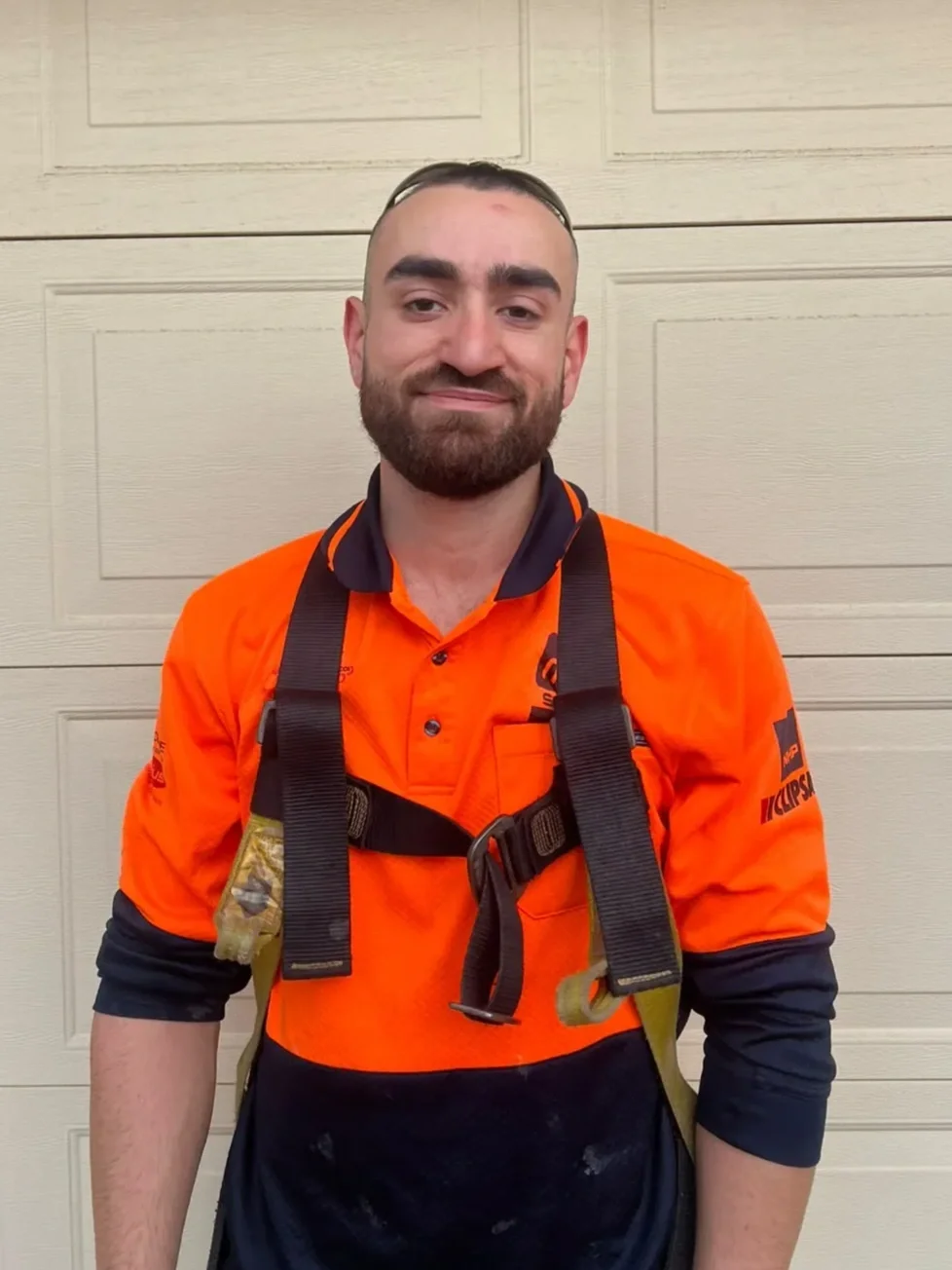 A man with a beard wearing an orange and black uniform, standing in front of a beige garage door.