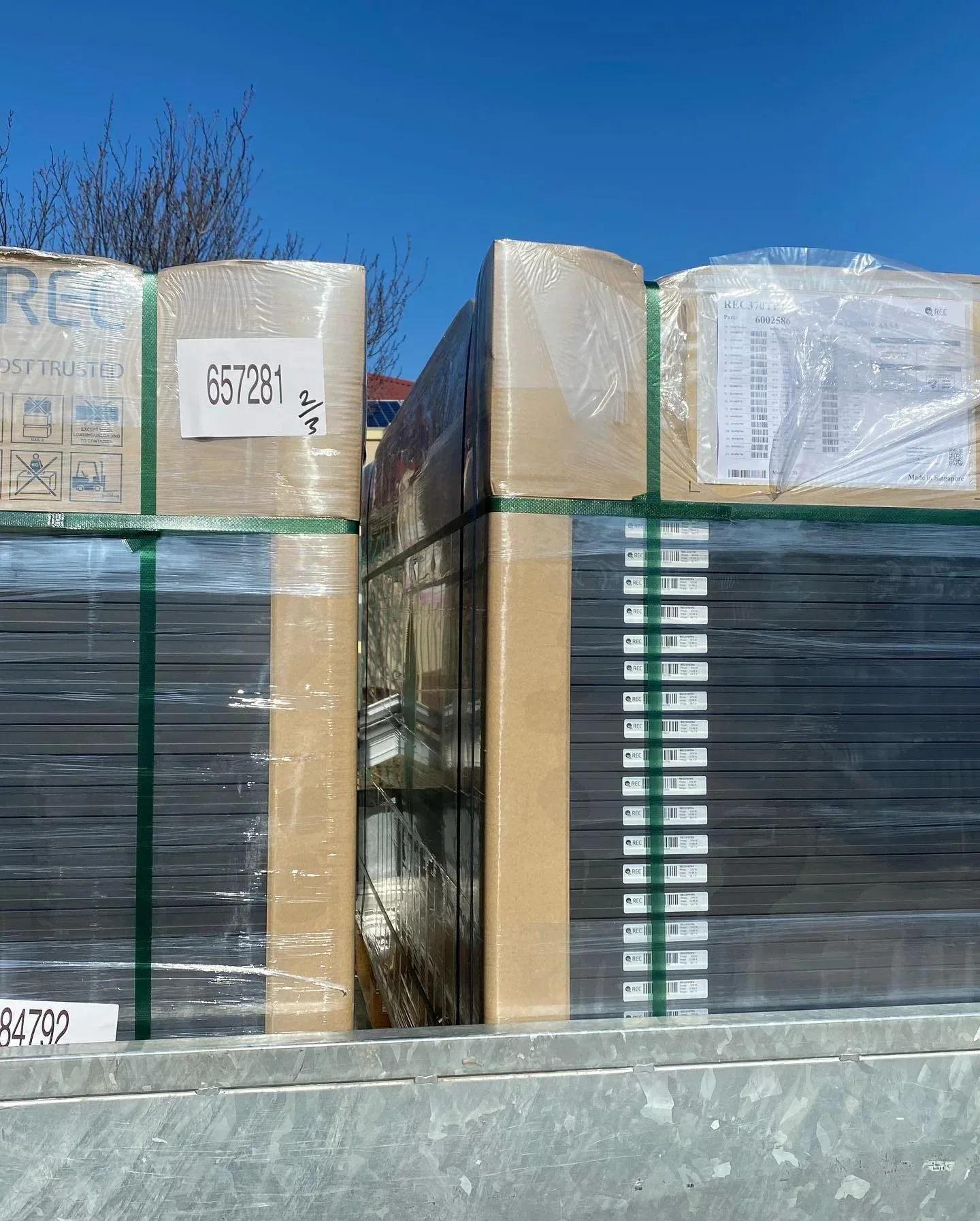Stacks of packaged goods wrapped in plastic and secured with green straps on a metal pallet outdoors against a backdrop of a blue sky and tree branches.