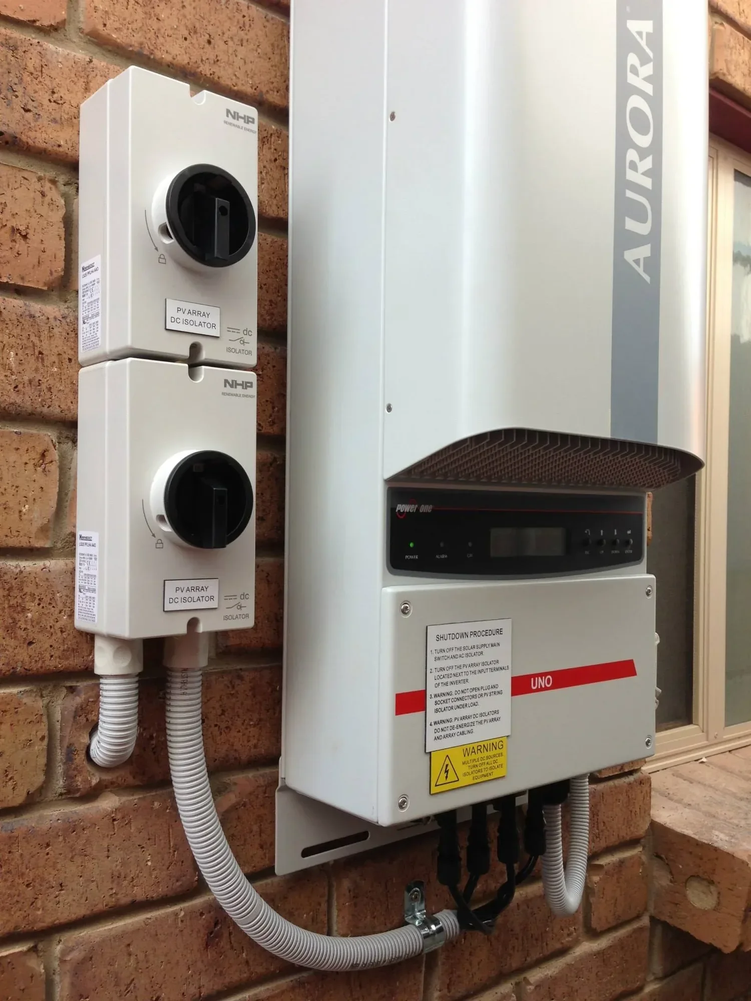 Electrical equipment mounted on a brick wall, including a solar inverter, PV array DC isolator switches, and associated wiring.