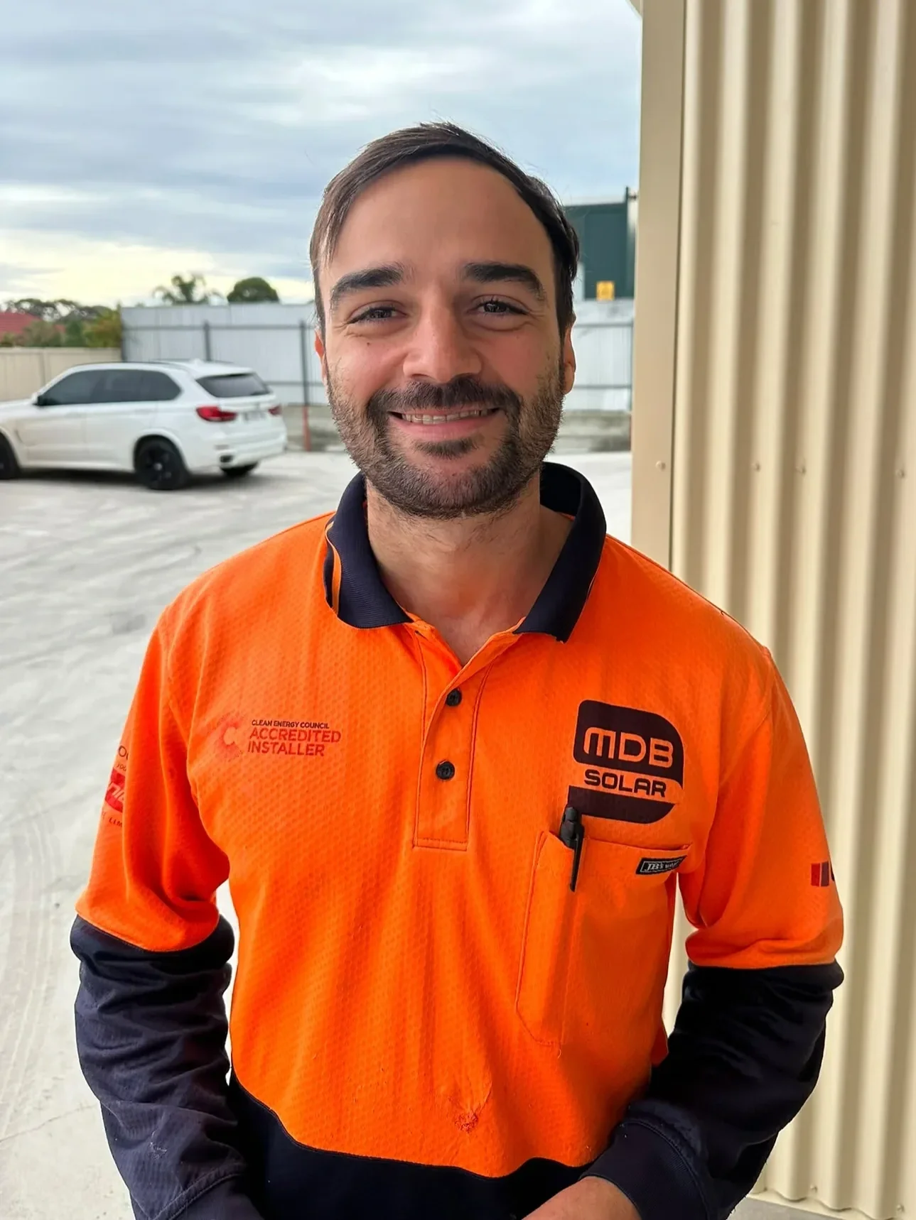 A man with short brown hair and a beard wearing an orange and black work uniform standing outdoors, smiling with a parking lot and cloudy sky in the background.