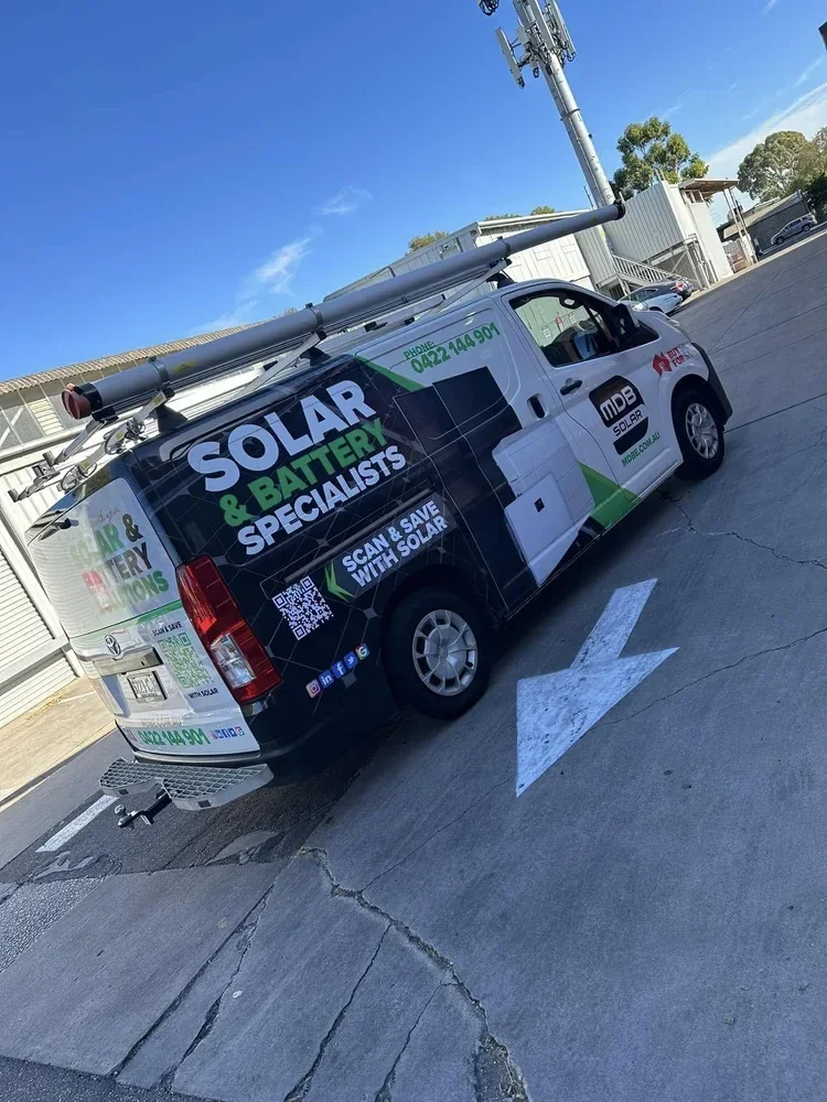 A company van for solar and battery specialists parked on a paved surface under a blue sky. The van has signage promoting solar panel installation and energy saving services, with a ladder on the roof and various contact details and social media icon