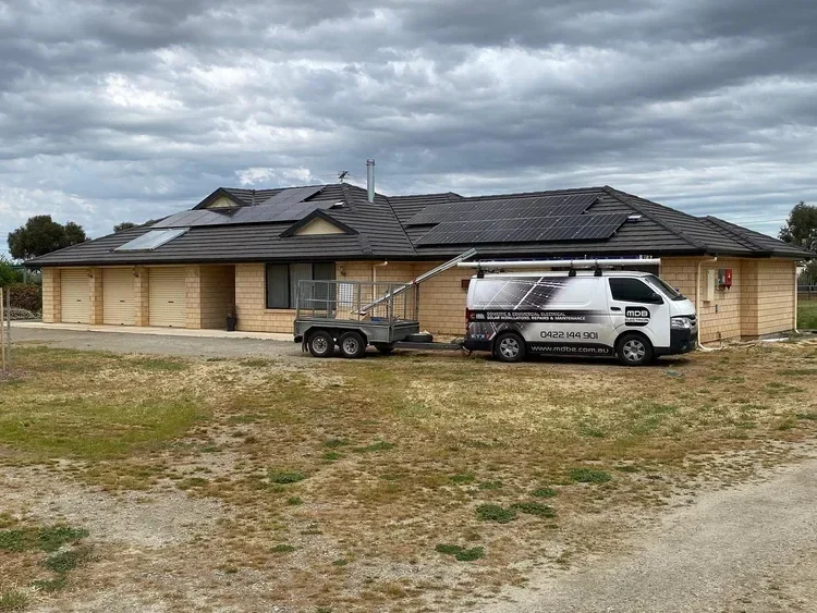 A house with solar panels on the roof, a white van with solar panel graphics parked in front, and a small trailer attached to the van, all on a patchy grass yard under a cloudy sky.