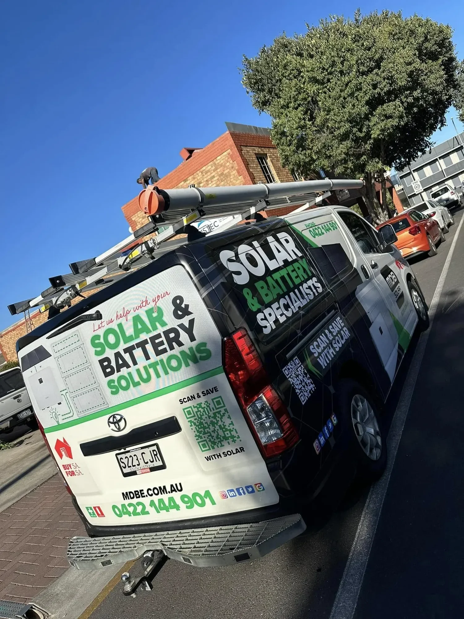 A white van with car battery and solar solutions advertisement, parked on a street next to a sidewalk and a large tree, with a brick building in the background and a man working on the roof.