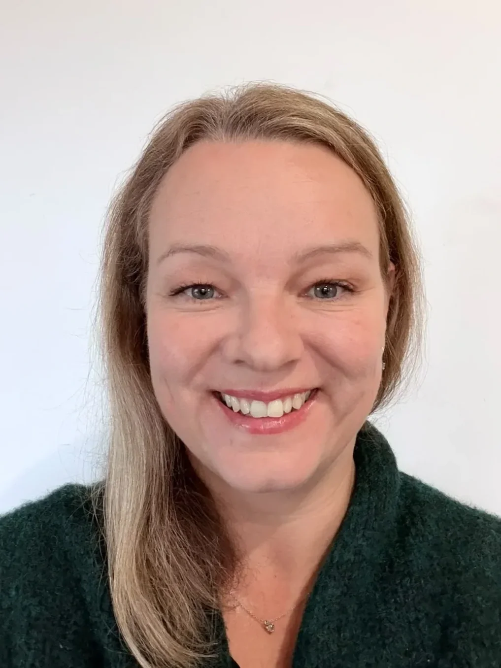 Close-up portrait of a smiling woman with light brown hair, blue eyes, wearing a dark green sweater and a small necklace, against a plain white background.