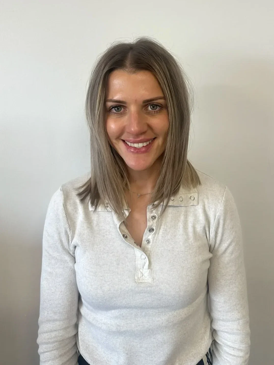 A woman with shoulder-length light brown hair, fair skin, and blue eyes is smiling at the camera. She is wearing a light beige, long-sleeved, ribbed shirt with metal studs on the shoulders against a plain white wall background.