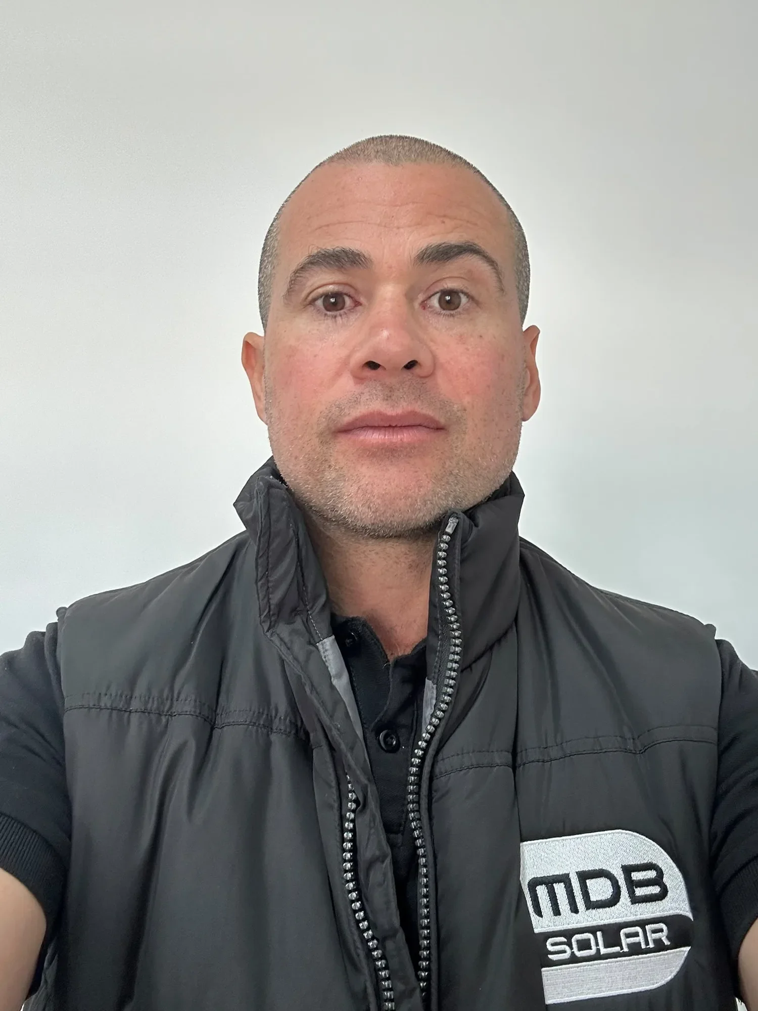 A man with short hair, wearing a black jacket with a high collar and a logo that reads 'MDP Solar' on the chest, takes a selfie against a plain light-colored background.