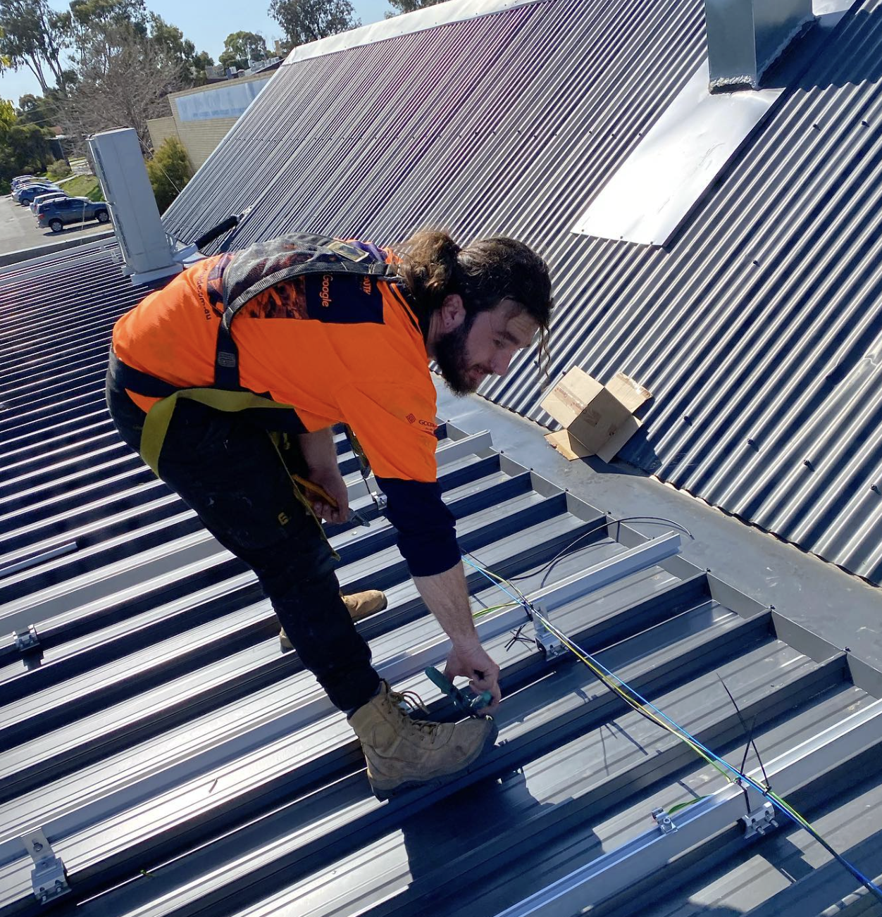 A man working on a metal rooftop, wearing an orange jacket, brown boots, and a safety harness, with tools and wires around him.