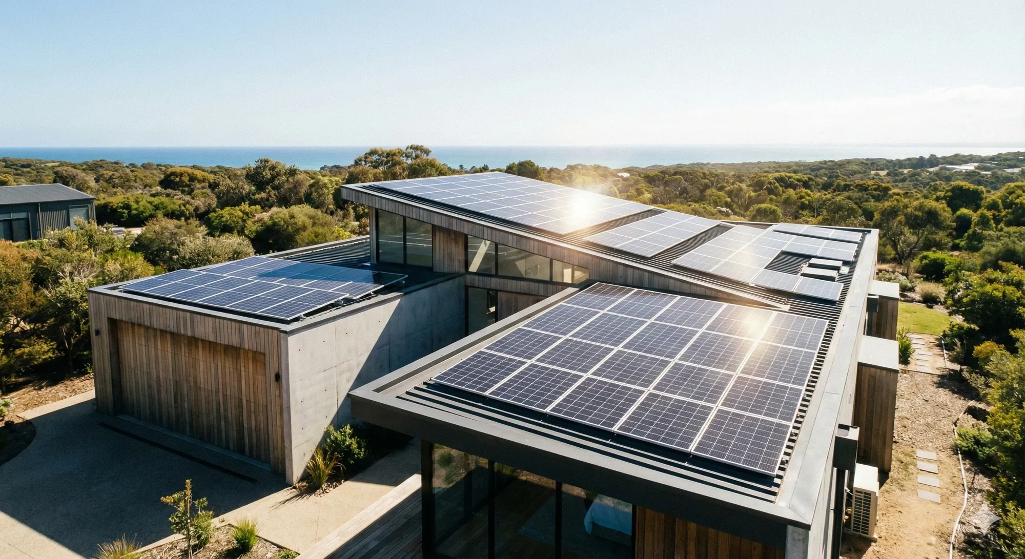 Modern house with flat roof covered in solar panels, situated among trees with a view of the ocean in the background.