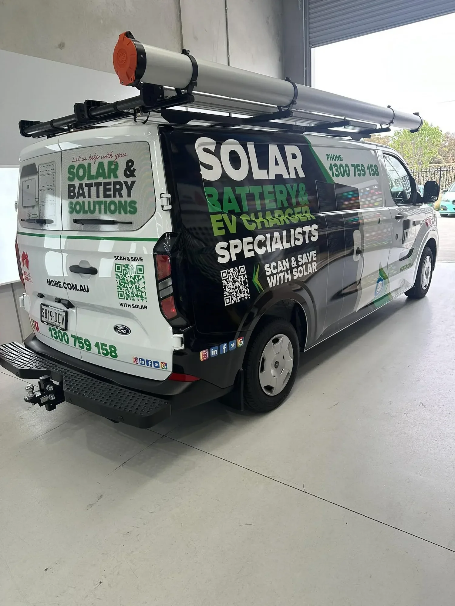 A white service van with colorful advertising for solar and battery solutions, parked indoors near an open garage door, with a ladder mounted on top.
