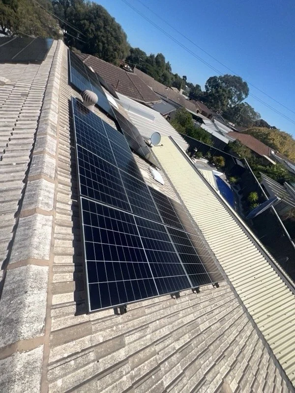 Solar panels installed on a residential rooftop with a clear blue sky and neighboring rooftops visible.