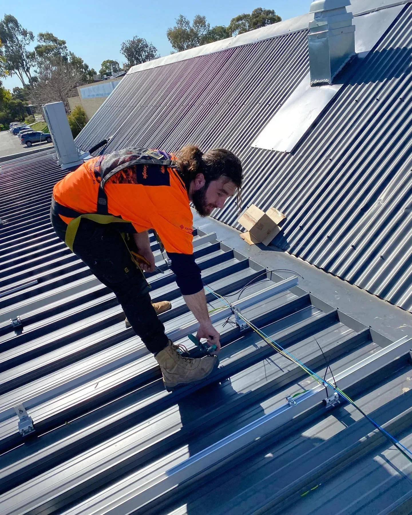 A man in an orange safety vest and tan work boots is installing or maintaining solar panels on a metal rooftop during the day. He is kneeling and working with wires and tools, with trees and parked cars visible below.