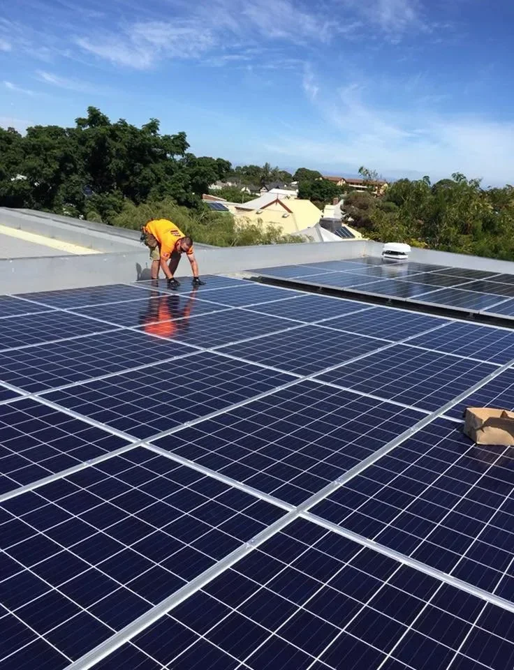 Worker installing solar panels on a rooftop during daytime with a blue sky and trees in the background.