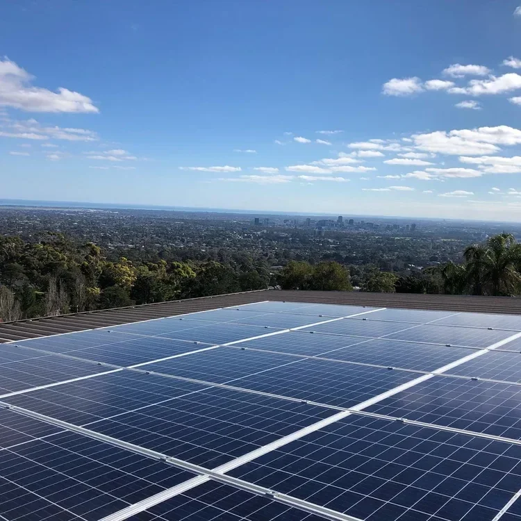 Solar panels on a rooftop with a city and forest in the background under a partly cloudy sky.