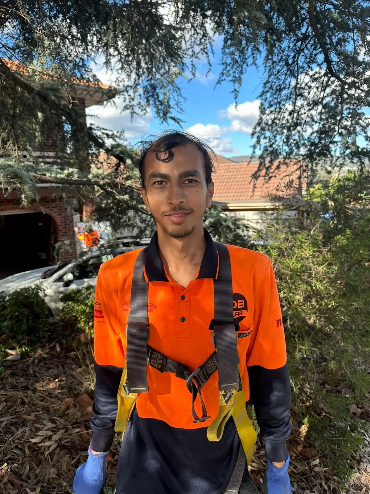 A young man with short, dark, curly hair and a thin beard, wearing an orange and black sports shirt with logos, has a camera strap around his neck and is standing outdoors in front of trees, bushes, and a house with a brick wall and a car in the background, under a partly cloudy sky.
