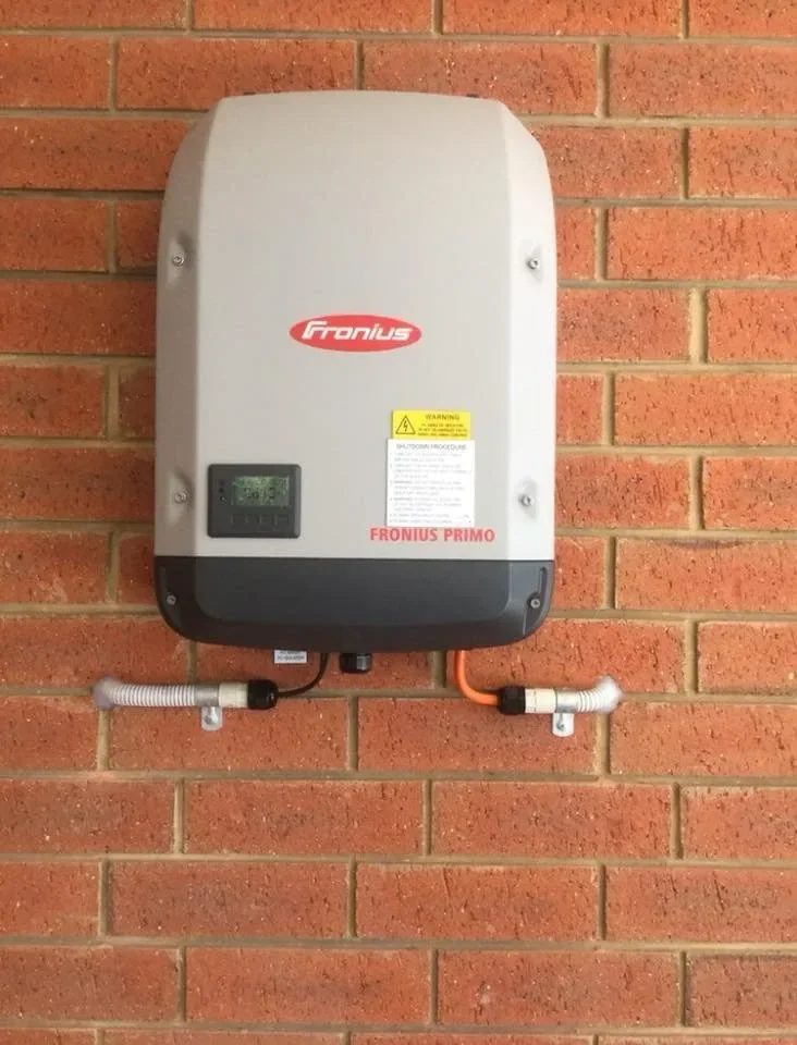Fronius Primo solar inverter mounted on a brick wall with electrical conduits connected at the bottom.