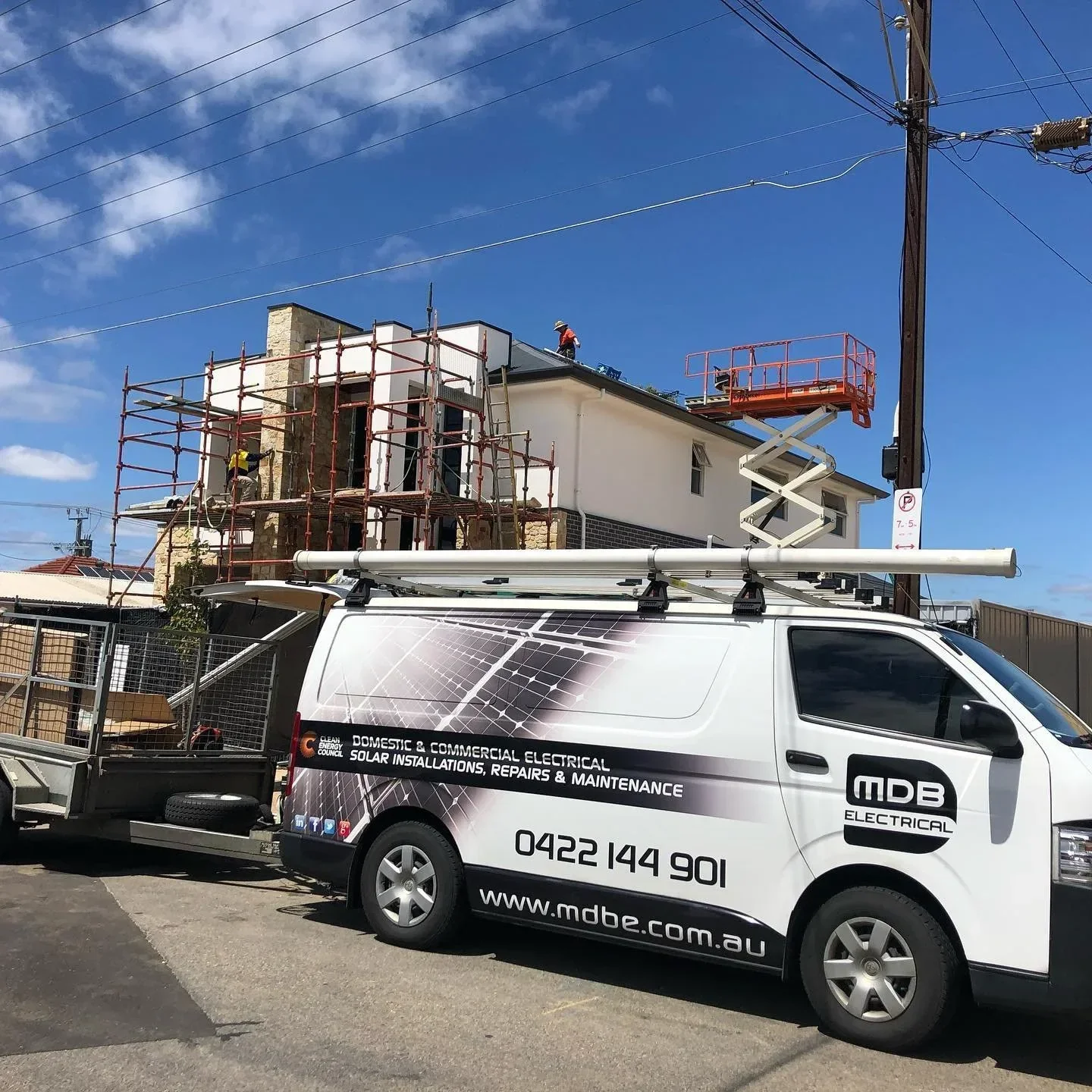Construction workers on scaffolding working on the roof of a house, with a lift and a utility pole nearby, in a residential area under a blue sky.