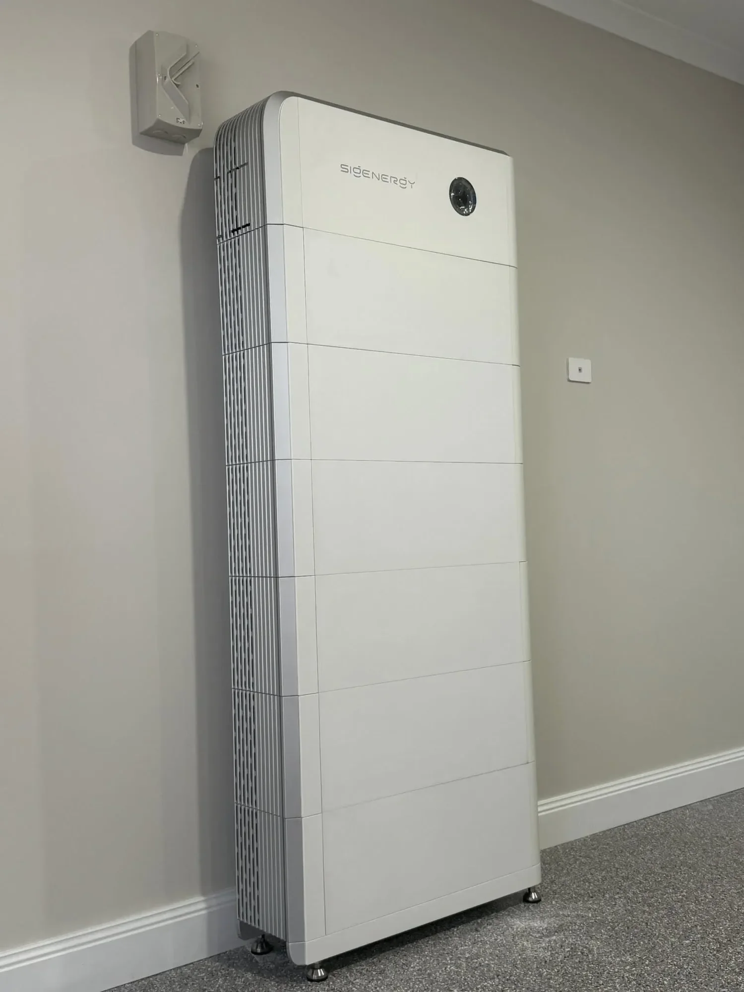 A white modern air purifier or heater with a circular display or sensor on the front, placed on a gray carpeted floor against a beige wall with a white baseboard. A small gray wall-mounted device is above it, and a white electrical outlet is on the r