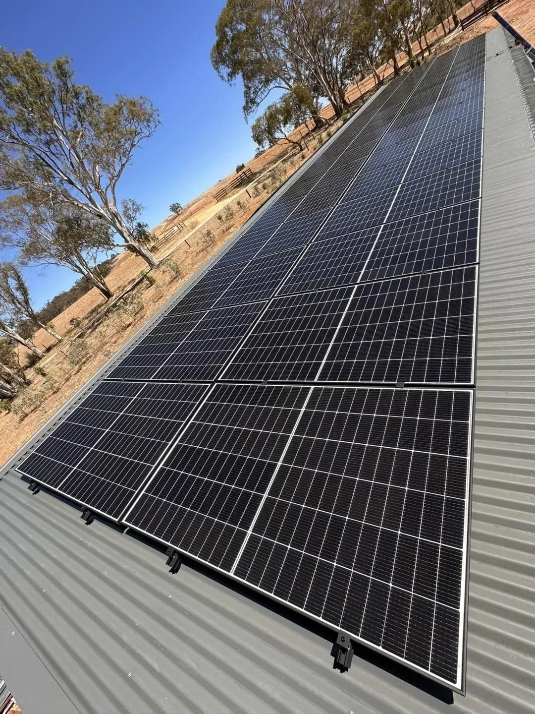 Solar panels installed on a metal roof, with trees and a clear blue sky in the background.
