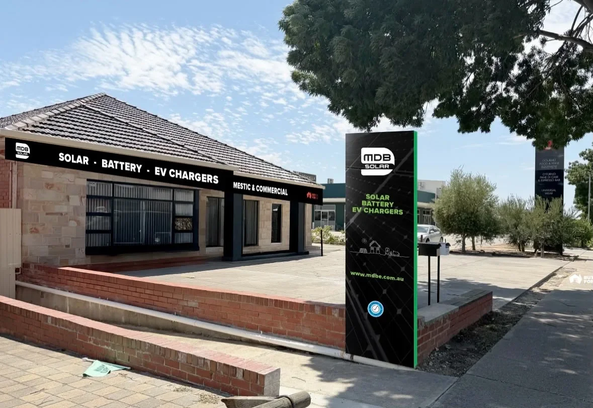 Storefront sign advertising solar, battery, and EV chargers with a black background and green accents on a tall signpost outside the store, which has a brick and stone exterior, large windows, and a black awning with white text.